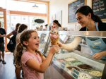 A smiling girl getting a double-scoop ice cream cone with sprinkles and a cherry from an employee in an ice cream shop