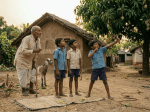 Boy sitting on a mat eating mangoes next to a goat and a sign that says Mango Challenge